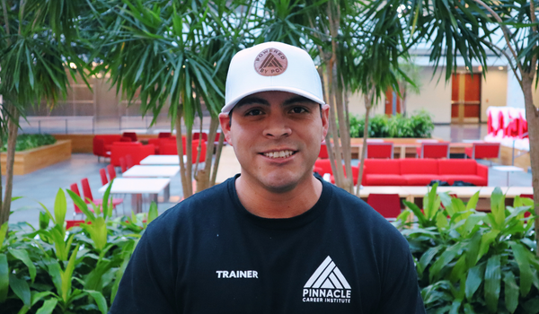 Edgar Gonzalez, GWO Instructor at Pinnacle Career Institute, wearing a PCI trainer shirt and hat, smiling in the atrium surrounded by greenery.