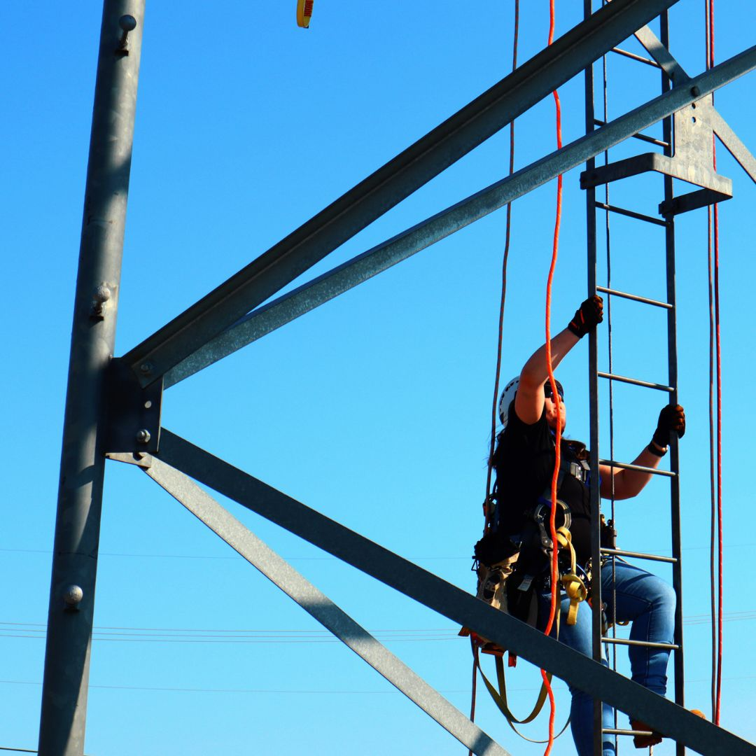 Student climbing a training tower ladder in full safety gear during PCI tower technician training.