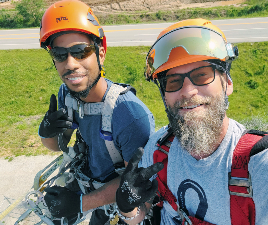 Two tower technician students in safety harnesses smiling during PCI’s hands-on tower climbing training.
