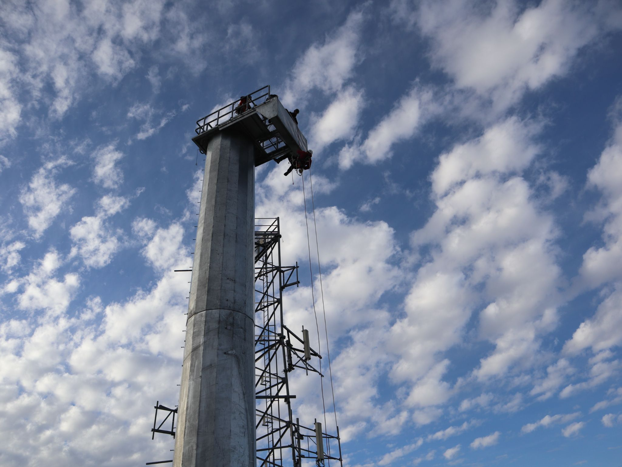 A tower technician scaling a tall communications tower under a partly cloudy sky during Pinnacle Career Institute training.