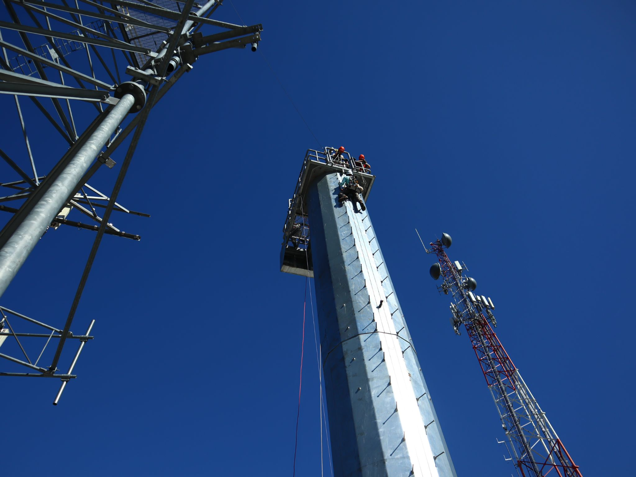 Tower technicians climbing a tall cell tower against a bright blue sky during hands-on training at Pinnacle Career Institute.